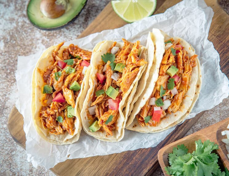 crockpot chicken tacos on a wood board with avocado, tomatoes and cilantro